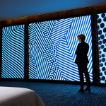 A sophisticated female hotel guest in modest, professional attire stands thoughtfully in a modern hotel room, observing a large, dynamic digital art installation projected onto a wall. The projection displays abstract, evolving patterns that subtly change color and form, creating an immersive, multi-sensory atmosphere. The room features minimalist, high-quality furnishings and soft, ambient lighting. The scene emphasizes an artistic, curated experience within a private space, with perfect anatomy, natural proportions, well-formed hands, and proper finger count. This is a professional photograph, high quality, safe for work, appropriate content, fully clothed, and family-friendly.